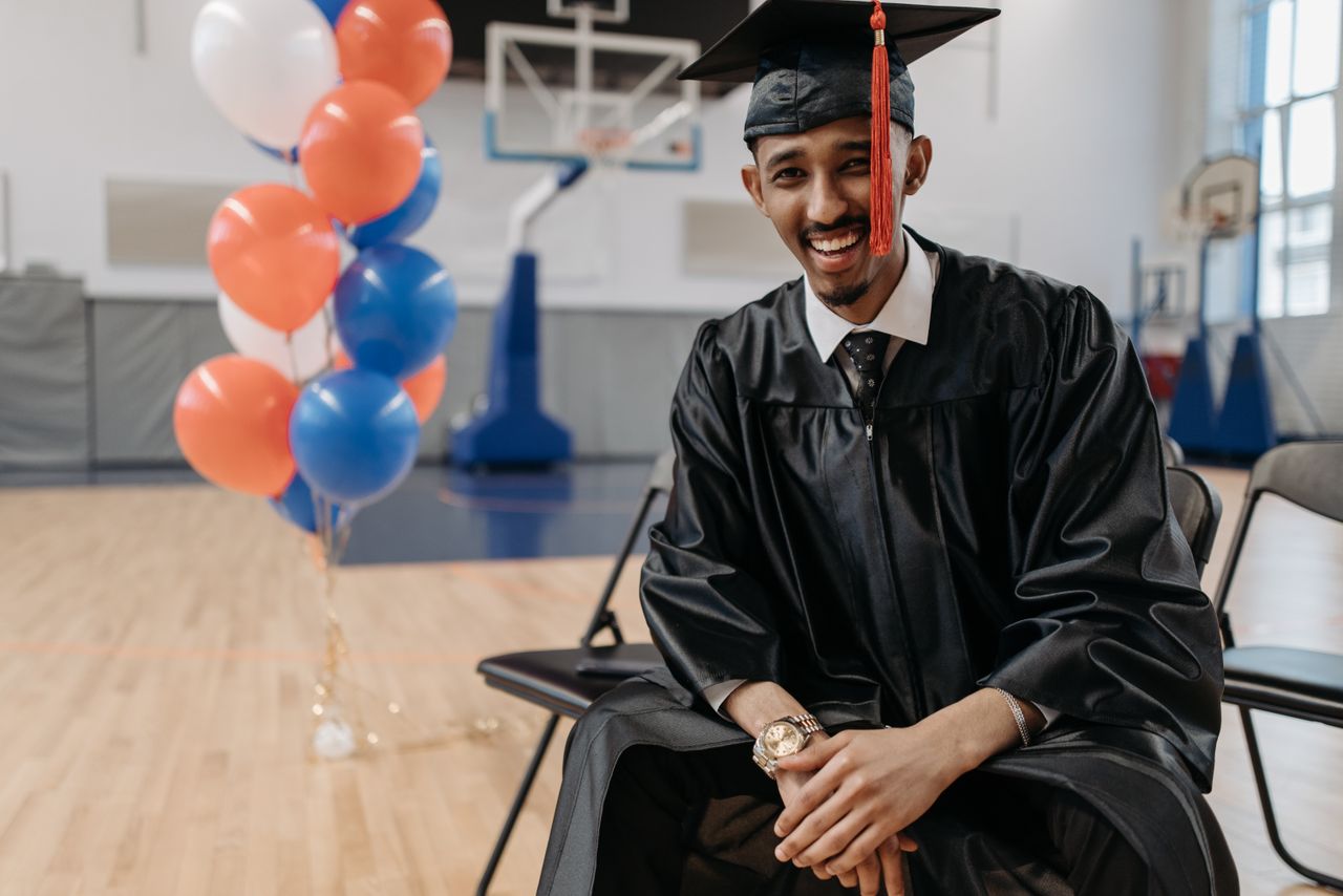 Services Smiling graduate sitting in a gymnasium with colorful balloons, celebrating his achievement.
