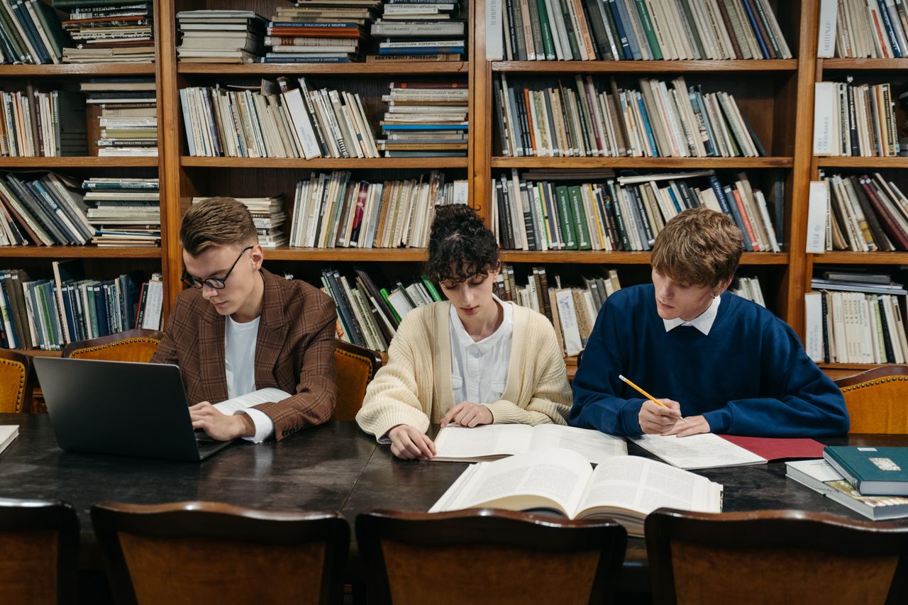 The Art of Drawing Readers In: Your attractive post title goes here Three students work on laptops and books in a university library filled with shelves.